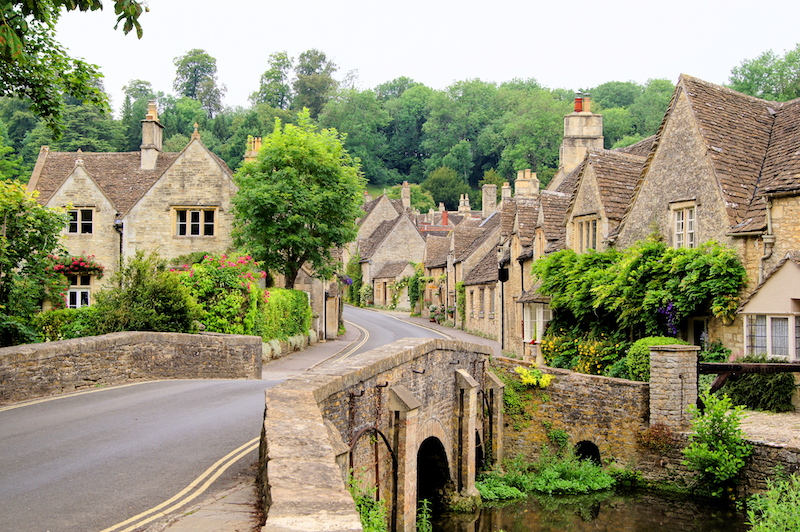 A beautiful Cotswolds village is shown, with traditional cottages shown over a bridge surrounded by trees.