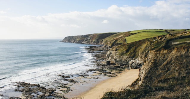 Coastline west of Porthleven on the south coast of Cornwall. Grassy cliff tops lead down to a rocky shoreline and beach. It is a sunny day and Penzance is a few miles beyond the headland.