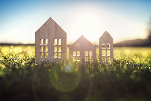 A wooden cut-out of a house sits in a green field, and sun is shining through the cut-outs for windows and doors.